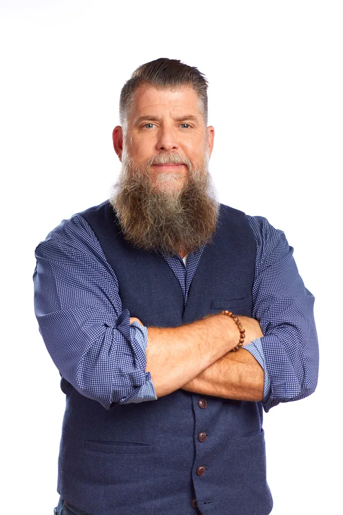 Studio portrait of a bearded man in a navy vest, arms crossed, on a white background
