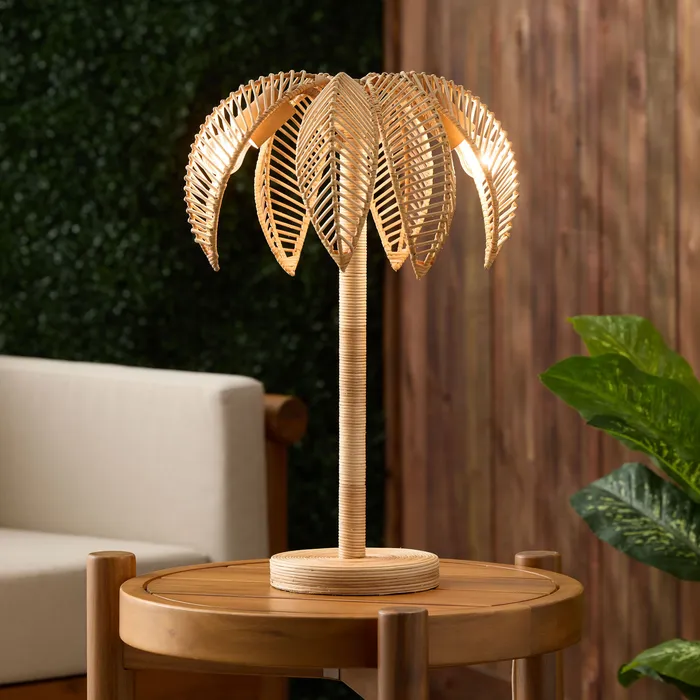 Woven rattan palm-leaf table lamp, lit, on a side table next to an outdoor sofa