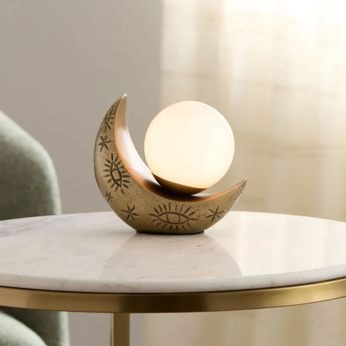 Brass crescent-moon table lamp with a glowing glass orb, on a marble side table